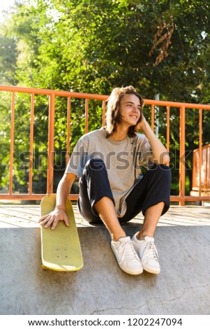 Photo of joyous skater boy 16-18 in casual wear sitting on ramp with skateboard in skate park during sunny summer day