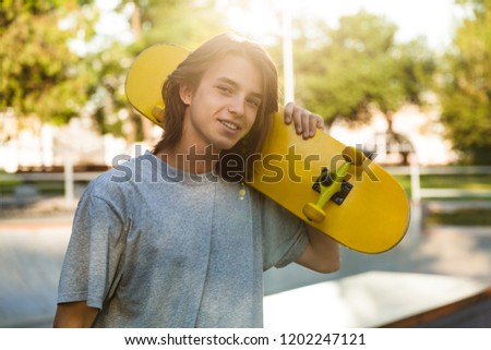 Photo of beautiful skater boy 16-18 in casual wear standing with skateboard in skate park during sunny summer day