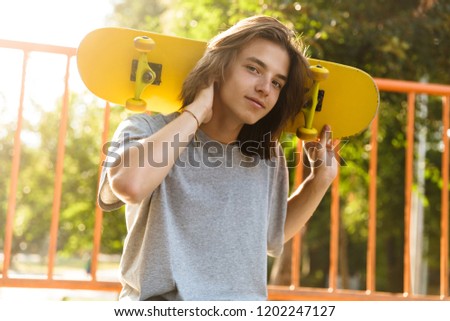 Photo of extreme guy 16-18 in casual wear sitting on ramp in skate park and holding skateboard over shoulders during sunny summer day