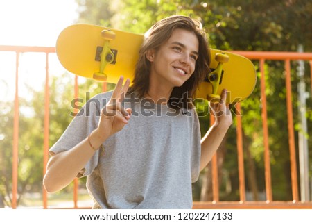 Photo of joyful guy 16-18 in casual wear sitting on ramp in skate park and holding skateboard over shoulders during sunny summer day