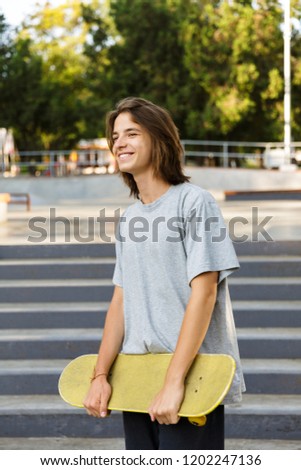 Photo of smiling skater boy 16-18 in casual wear standing with skateboard in skate park during sunny summer day