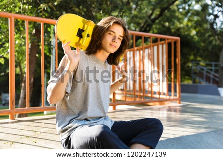 Photo of teen guy 16-18 in casual wear sitting on ramp in skate park and holding skateboard over shoulders during sunny summer day
