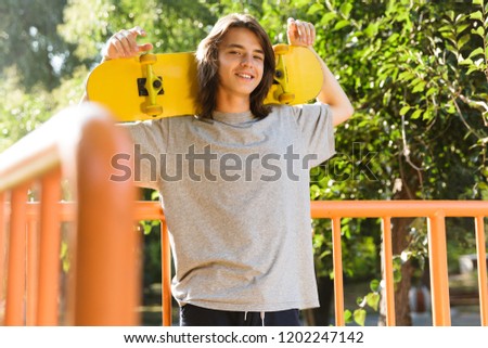 Photo of young guy 16-18 in casual wear standing in skate park and holding skateboard over shoulders during sunny summer day