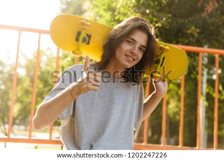 Photo of cheerful guy 16-18 in casual wear sitting on ramp in skate park and holding skateboard over shoulders during sunny summer day