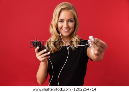 Image of young excited woman using phone listening music gives you one earphone isolated over red background.
