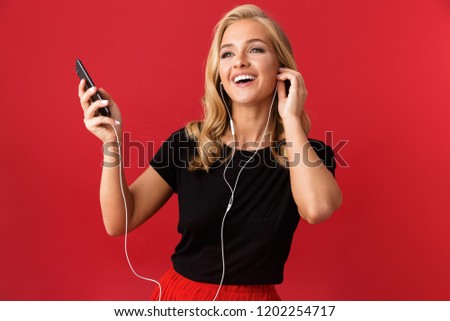 Image of young excited woman using phone listening music with earphones isolated over red background.