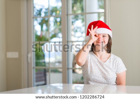 Down syndrome woman at home wearing christmas hat with happy face smiling doing ok sign with hand on eye looking through fingers
