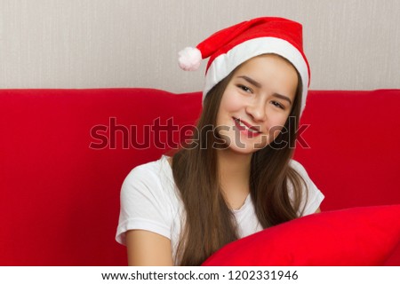 Christmas mood. A young European girl with long dark hair in a Santa Claus hat is sitting on the sofa and smiling.