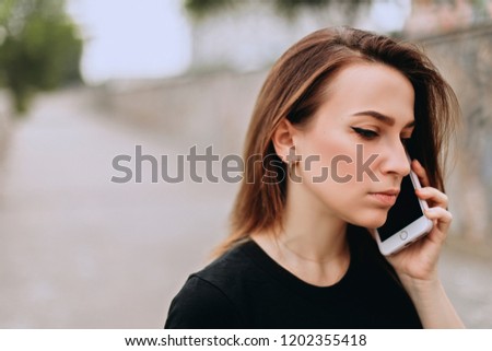 Photo of serious modern woman walking on summer day and talking on smartphone. Girl wearing black t-shirt. Brunette short hair.