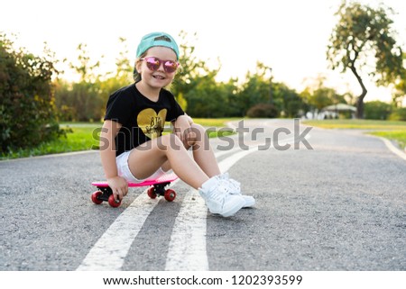 Fashion little girl child sitting on skateboard in city, wearing a sunglasses and t-shirt