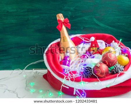 golden champagne bottle in red santa gift bag with lights and balls on green background with sopy space. merry christmas and happy new year concept