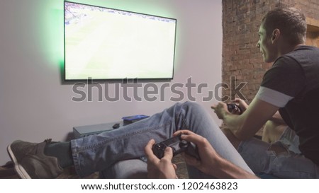 Two men playing video game Pro Evolution Soccer. Hands holding console controller. Football or soccer game on the television. Widescreen tv hang on the wall Royalty-Free Stock Photo #1202463823