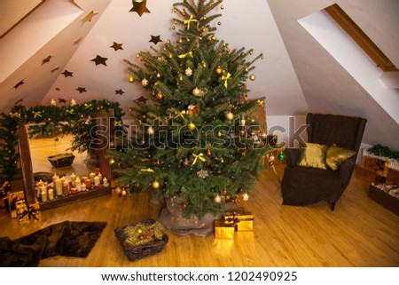 Christmas decorations in the studio, big natural fir tree with golden balls, bows and snowflakes, wooden fireplace and candles on it and brown cozy armchair