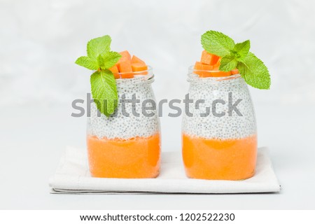 Chia seeds pudding with pumpkin puree in beautiful glasses with green mint leaves and cut fresh ripe orange vegetable on light gray background - raw vegetarian sweet organic dessert.