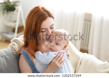 Lovely sweet morning of cute young mother cuddling her baby boy at couch in living room, kissing him on forehead. Family bonds, maternity leave, childhood, parenting and motherhood concept