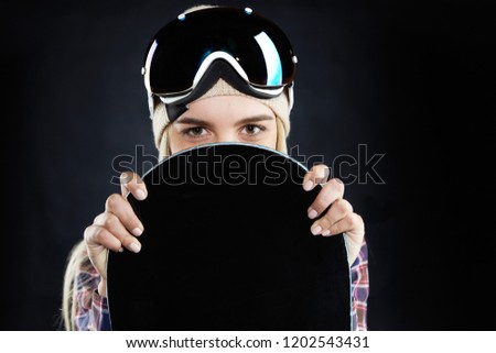 People, travel, recreation and extreme sports concept. Portrait of mysterious positive young woman snowboarder with protective goggles on her head, hiding behind black board and looking at camera