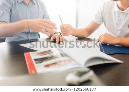 Picture of two men sit at table together. They hold hands on documents. Guy on right gives pen to man on left. There is journal on table. Small white car is in front of picture.