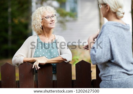 Two mature female neighbours talking through fence about everyday life stuff Royalty-Free Stock Photo #1202627500