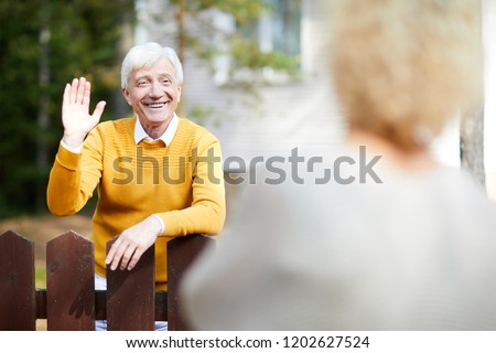 Cheerful and friendly mature man waving hand to his neighbour while standing by fence Royalty-Free Stock Photo #1202627524