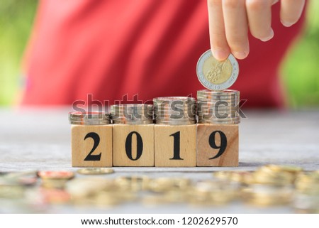 Asian kid's hand putting coins to stack of coins on wooden block with text 2019. Concept of money saving, financial, new year resolution.