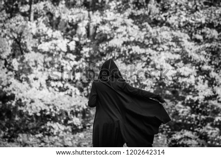 View from the back on a black witch in a mantle stays in a forest. Black and white moody photography. The concept of Halloween and witchcraft.