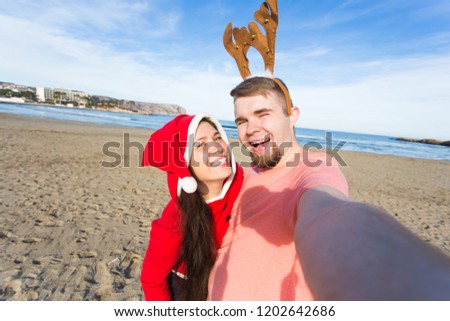 Holidays, people and Christmas concept - couple wearing christmas costumes taking selfie photo at sandy beach