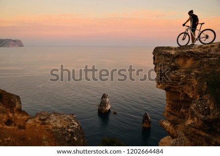 Cyclist with a bicycle stands on the edge of a cliff on the background of the sea at sunset