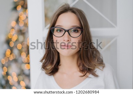 Headshot of lovely young female model in transparent glasses, has dark hair, dressed in casual clothes, enjoys domestic atmosphere, poses against New Year tree. People and lifestyle concept.