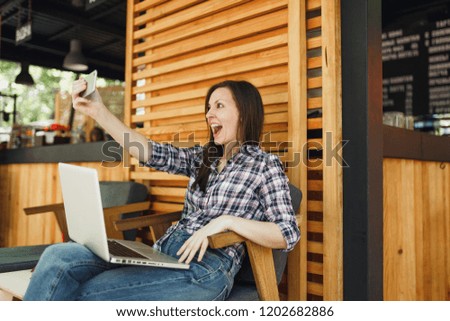 Woman in wooden outdoors street summer coffee shop sitting with laptop pc computer, doing selfie shot on mobile phone, relaxing during free time. Mobile Office. Lifestyle freelance business concept