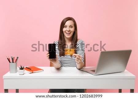 Excited woman holding mobile phone with blank empty screen, credit card sit work at white desk with contemporary pc laptop isolated on pastel pink background. Achievement business career. Copy space