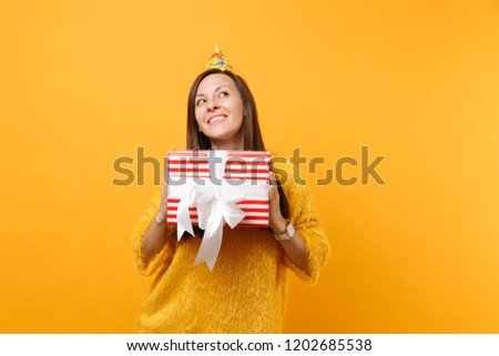 Dreamy young woman in birthday hat looking up, holding red box with gift, present enjoying holiday isolated on bright yellow background. People sincere emotions, lifestyle concept. Advertising area