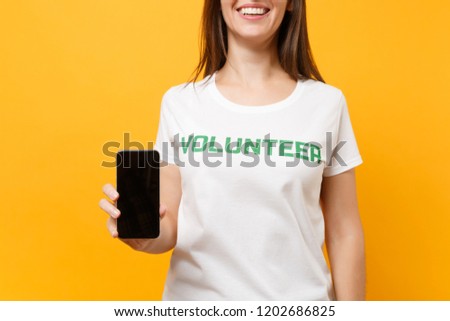 Woman in white t-shirt written inscription green title volunteer hold mobile phone with blank empty screen isolated on yellow background. Voluntary free assistance help, charity grace work concept