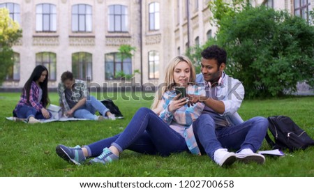 Young couple sitting on lawn on campus and watching pictures on smartphone