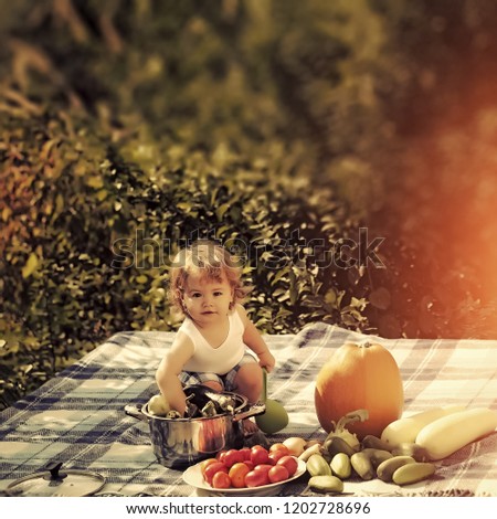 One little smiling boy at picnic sitting with ladle pot orange pumpkin squash and cucumber red tomato on checkered plaid looking forward on natural background sunny day, square picture