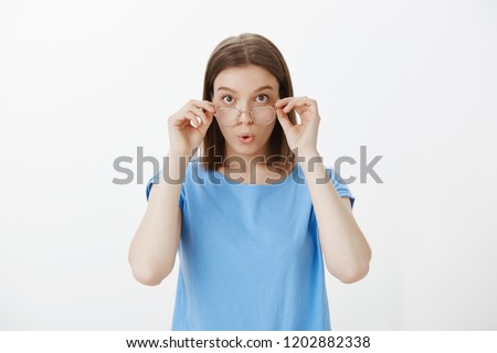 Wow, give me two. Portrait of impressed interested attractive woman in nerdy glasses, touching rim of eyewear with both hands, folding lips, being curious while picking new book in store