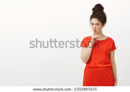Strict good-looking Caucasian female with curly hair in casual red dress, saying shh making warning shush gesture with index finger over mouth, frowning, wanting silence, asking keep voice down