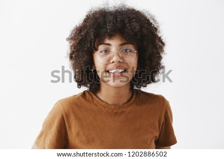 Close-up shot of friendly good-looking african american female student in transparent glasses and brown t-shirt smiling with joyful pleasant smile being satisfied with how things going over gray wall