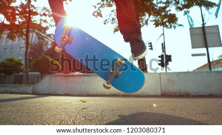 CLOSE UP, LENS FLARE: Unrecognizable male athlete does a fakie trick with his blue skateboard on sunny day at the park. Young skater rolling down the pavement jumps and does a flip with his deck.