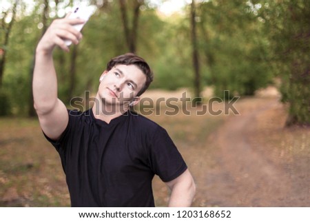 Attractive young caucasian man with dark hair making selfie in park. Outdoors, autumn day, golden leaves on the groung, green trees. Copy space
