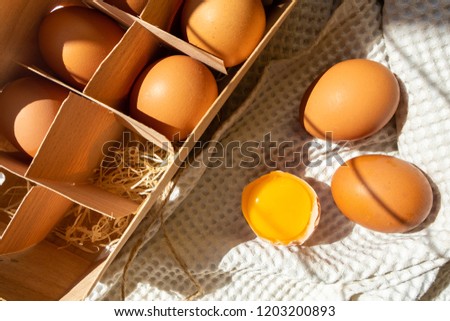 Chicken eggs in the box with half broken egg with yolk on the white towel in sun rays.