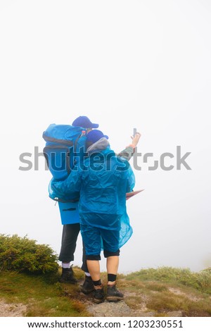 Couple man and woman in blue raincoat standing at the cliff in mountains taking selfie. Travellers hikers in boots and blue hats taking pictures at cliff in mountains in mist. Man holds phone in hand