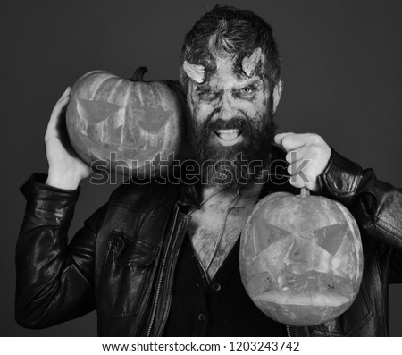 Devil or monster with October decorations. Man wearing scary makeup holds pumpkins on bloody red background. Demon with horns and mad face holds carved jack o lanterns. Halloween party concept