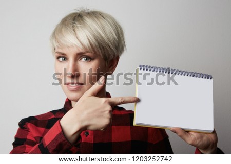 Portrait of attractive blonde woman posing in a red shirt holding white empty notepad and poiting hang.Mock-up.White background