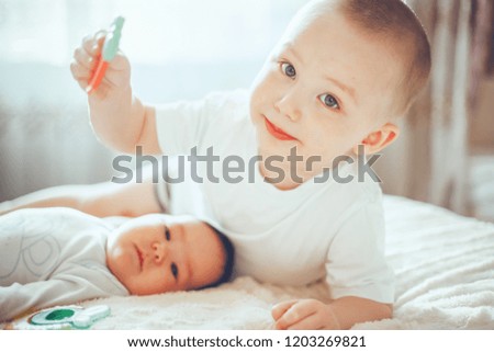 elder brother blonde boy son playing with little baby sister with black hair on bed at home children in white clothes