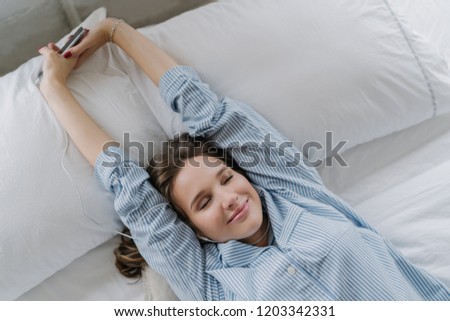 Satisfied woman stretches in bed, dressed in nightclothes, closes eyes with pleasure, holds cell phone, listens music in earphones, recreats in bedroom. People, good rest and modern technologies