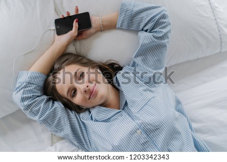 Relaxation concept. Pretty girl with pleased satisfied expression, dressed in pyjamas, rests in bed, holds smart phone, listens pleasant music in earphones. View from above. Woman has good day at home