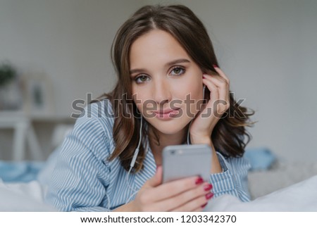 Close up shot of attractive brunette young woman downloads songs from network, uses smart phone for entertainment, dressed in nightclothes, rests in bed, has lazy day, looks seriously at camera