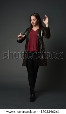 full length portrait of brunette girl wearing long leather coat. standing pose on grey studio background.