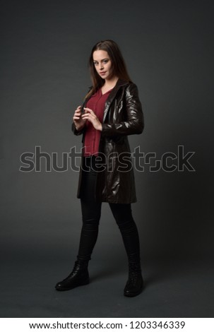 full length portrait of brunette girl wearing long leather coat. standing pose on grey studio background.