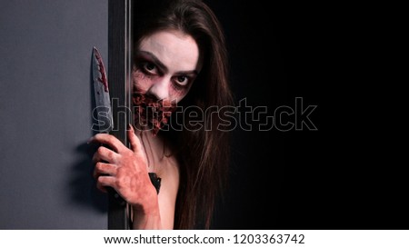 Makeup for halloween party. A girl with a bloodied knife in her hand looks out from behind a gray wall. Imitation of a bloody wound and a wired mouth. Black background. Copy space.